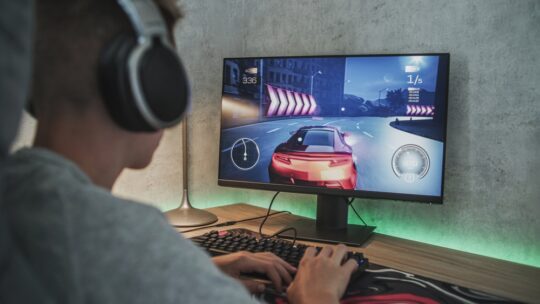 teenager wearing headphones plays online car video game on personal computer. Professional young gamer spends time at home, enjoys gameplay in street racing drift simulator, uses keyboard.